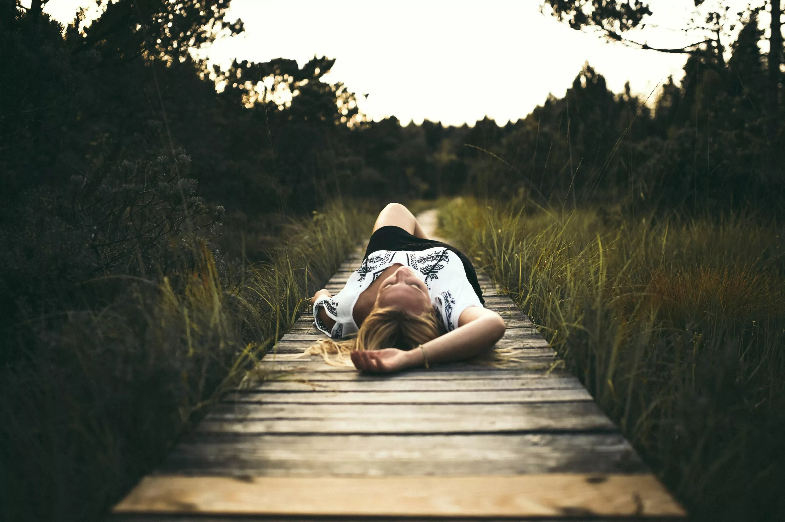 A woman lying on her back on a wooden boardwalk through long grass, looking up at the sky