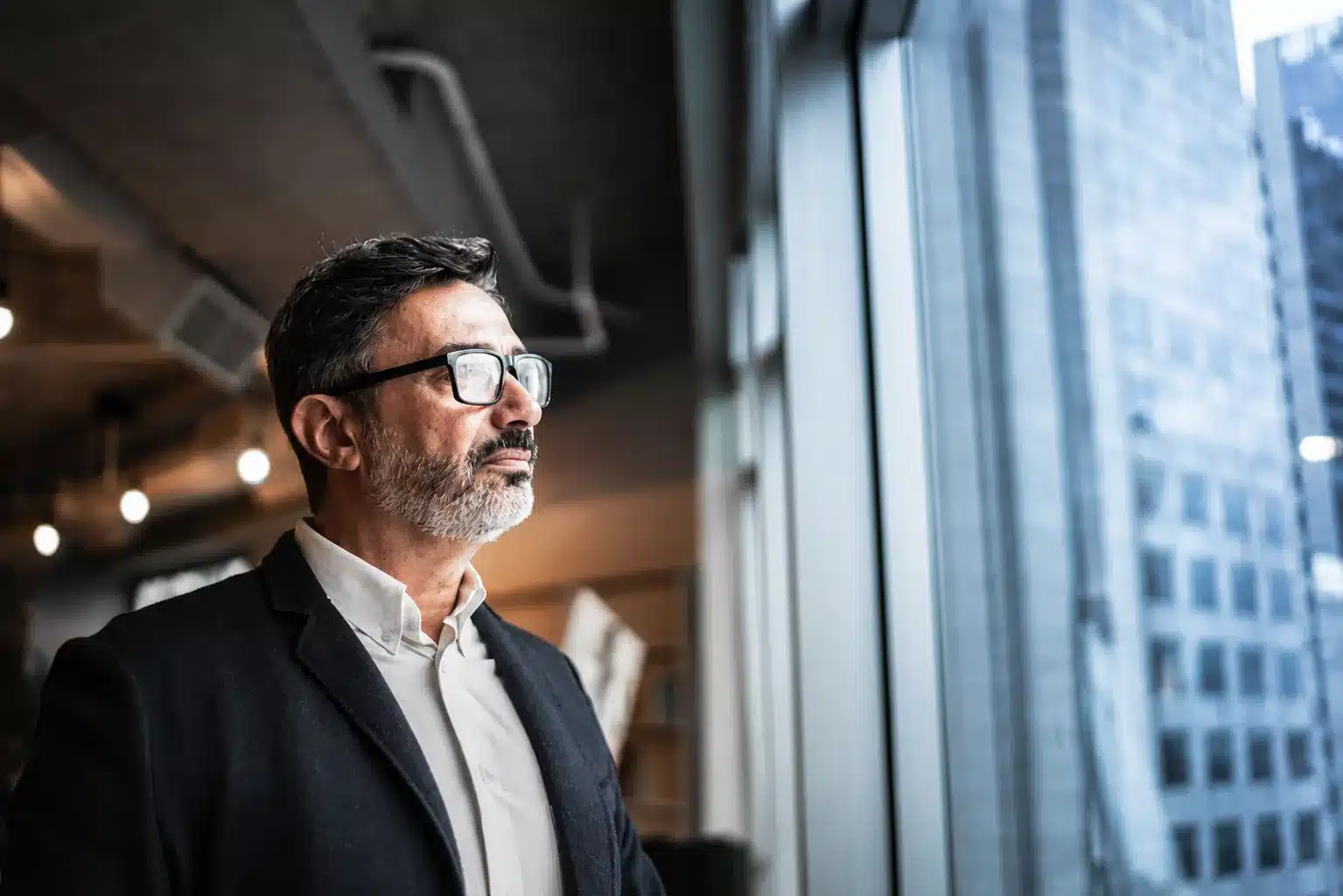 A senior leader in glasses and a dark jacket standing at a tall office window, looking out over the city while he thinks