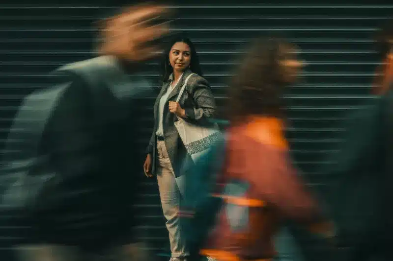 A woman in a grey blazer standing still and composed on a city street while colleagues blur past her