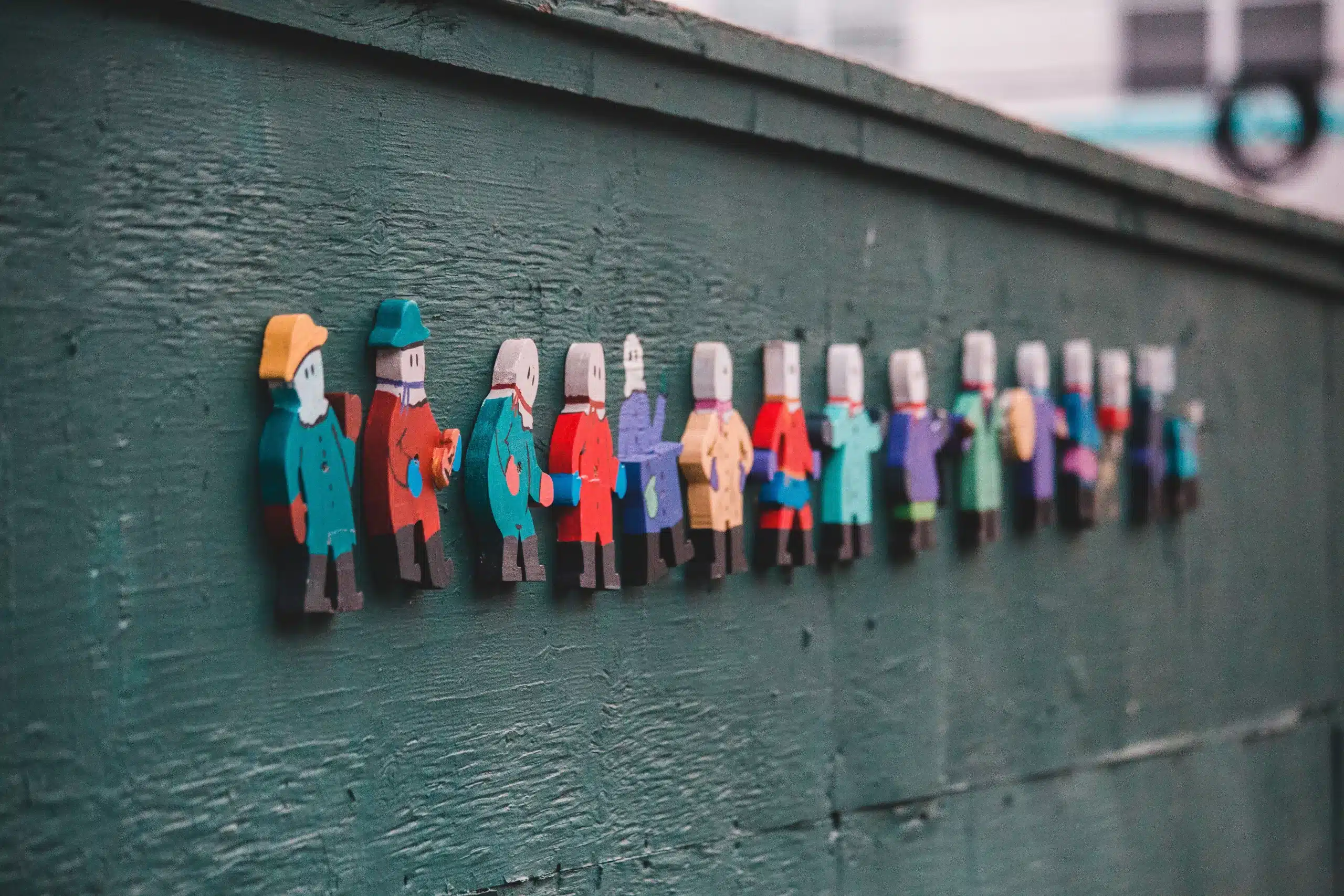 A row of small painted wooden figures in different coloured coats fixed in a line along a green wooden wall
