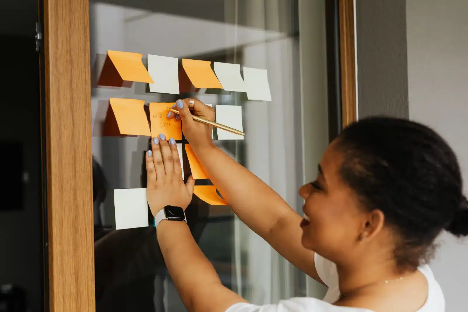 A smiling woman writing on orange and white sticky notes arranged on a glass window, putting her reminders where she can see them.