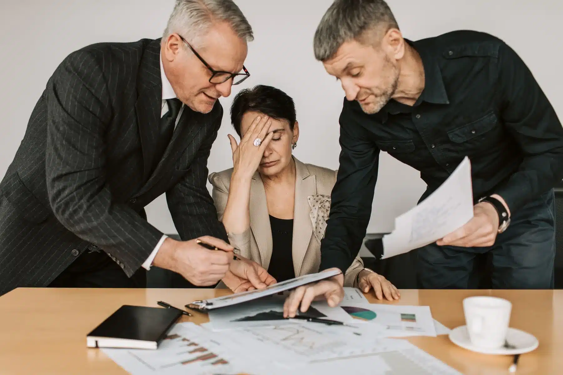 A woman at a meeting table with her hand to her forehead while two older colleagues lean over her with papers.