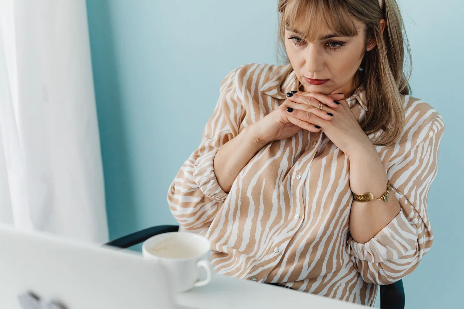 A woman at a desk with her hands clasped under her chin, looking at an open laptop without yet beginning.