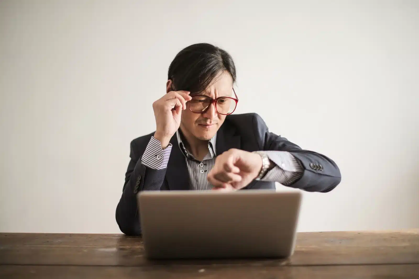A man in a suit at a desk with a laptop, adjusting his glasses as he glances at his wristwatch, suddenly aware of the time.