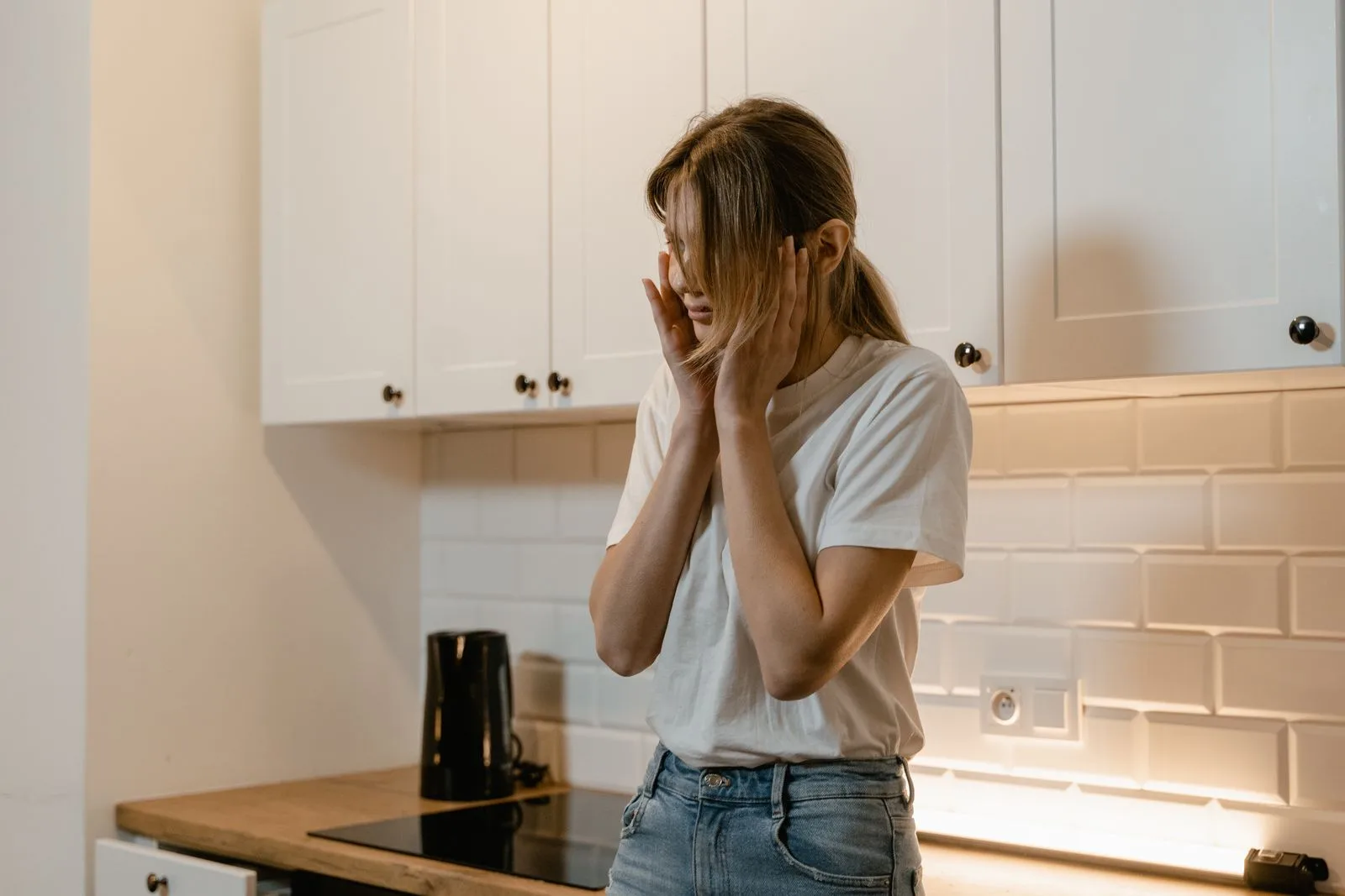 A woman standing in a kitchen with her hands to her face, appearing overwhelmed.