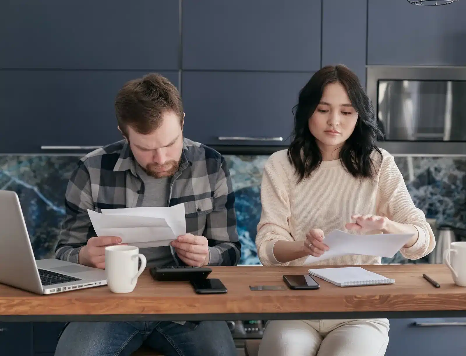 A man and a woman sitting side by side at a kitchen table, quietly reading through papers.