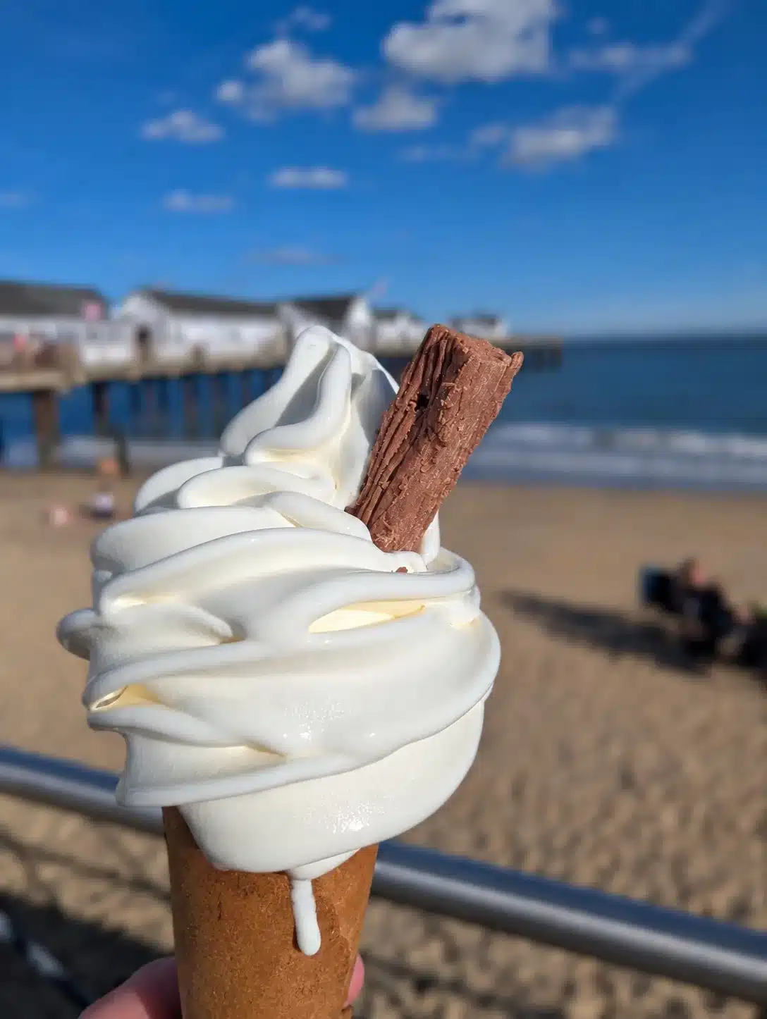 A soft serve ice cream with a chocolate flake in focus, with a softly blurred sandy beach, sea and pier beneath a blue sky behind it.