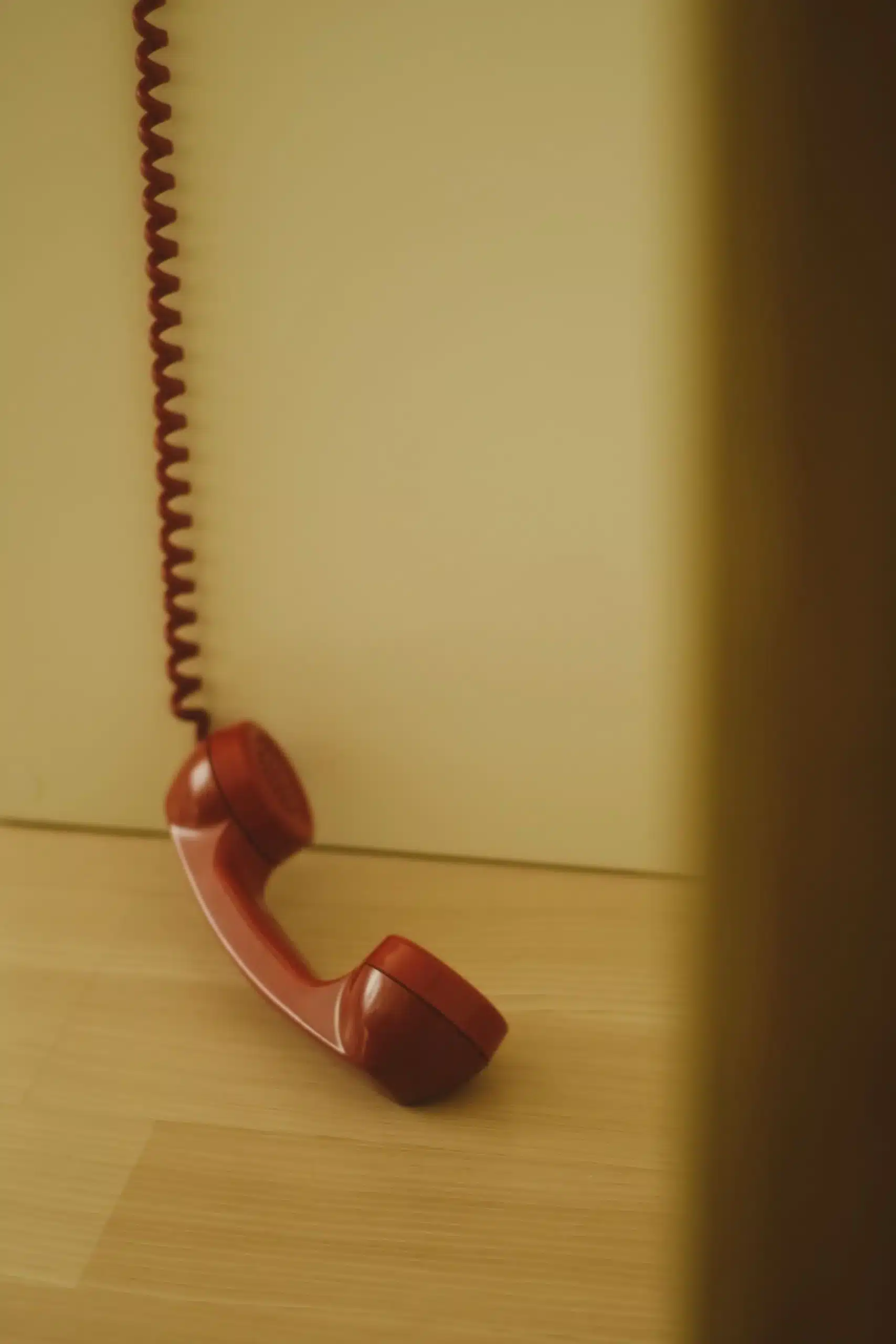 A red telephone receiver left off the hook, with its coiled cord hanging down towards a wooden floor in muted warm light.