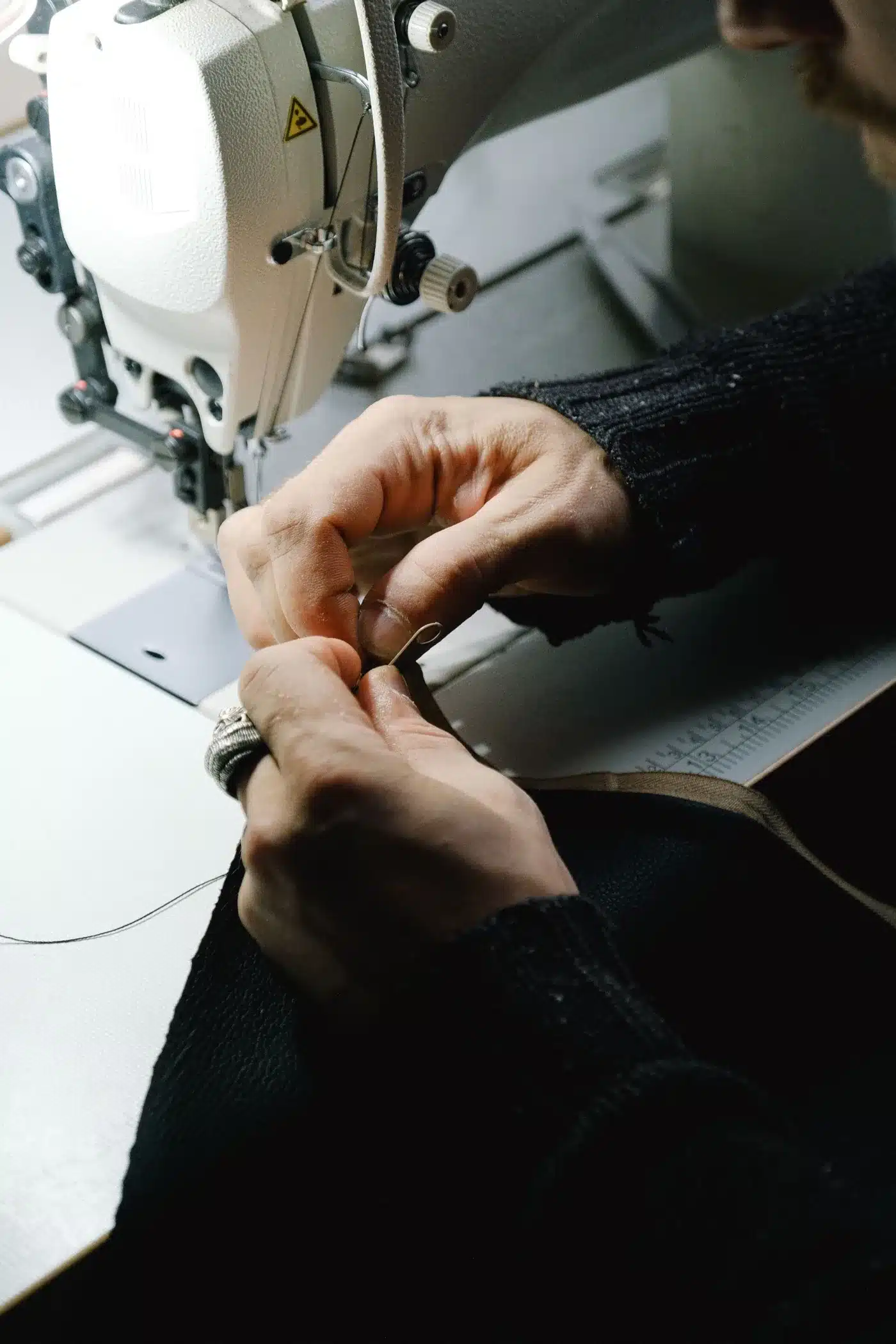 Hands threading a needle beside an industrial sewing machine in a workshop.