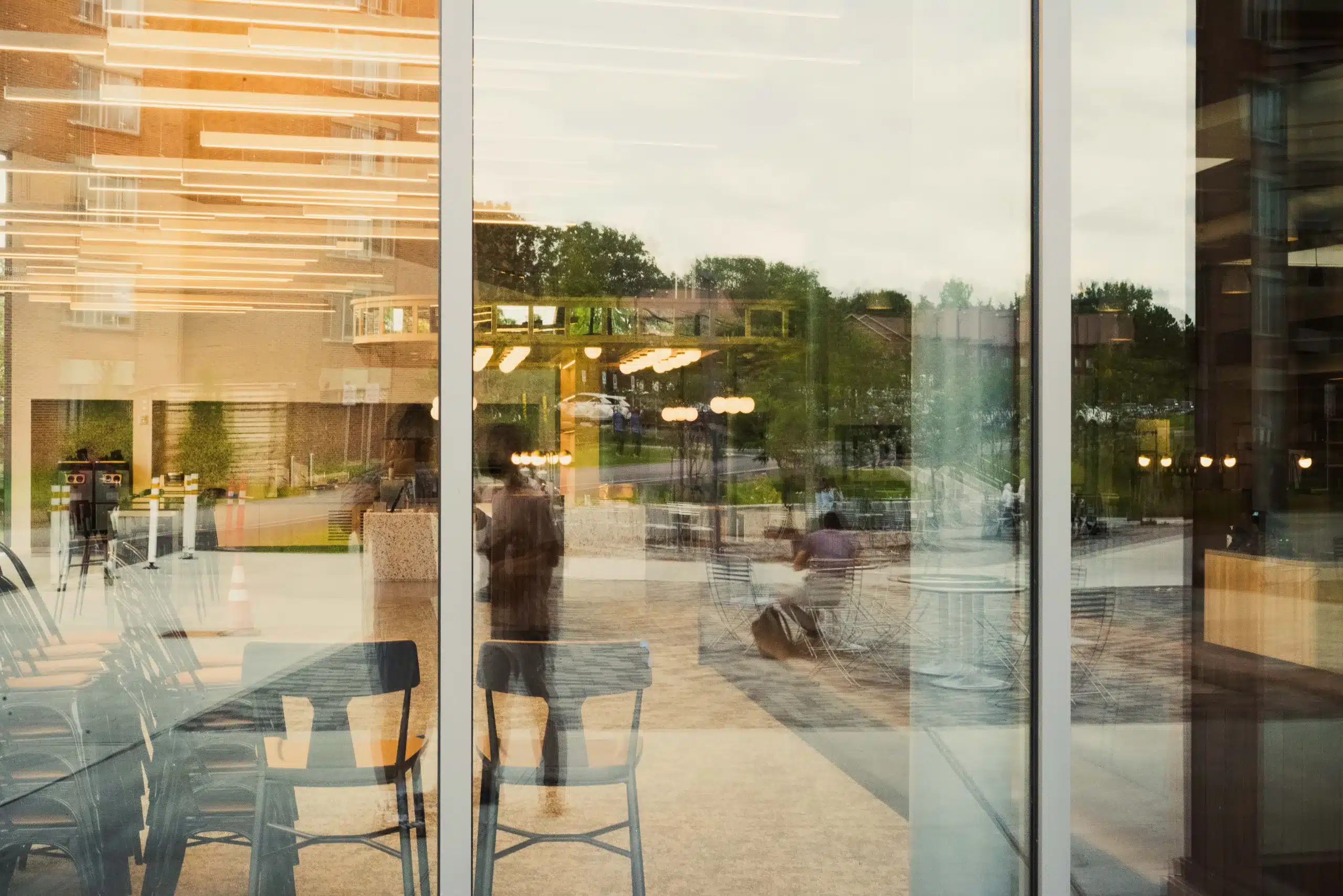 Café chairs and tables seen through large windows, with warm indoor light and reflections of trees and people outside.
