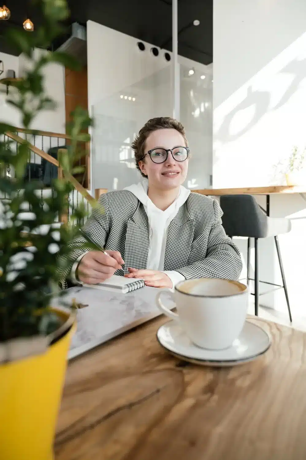 A smiling person wearing glasses writes in a notebook at a café table.