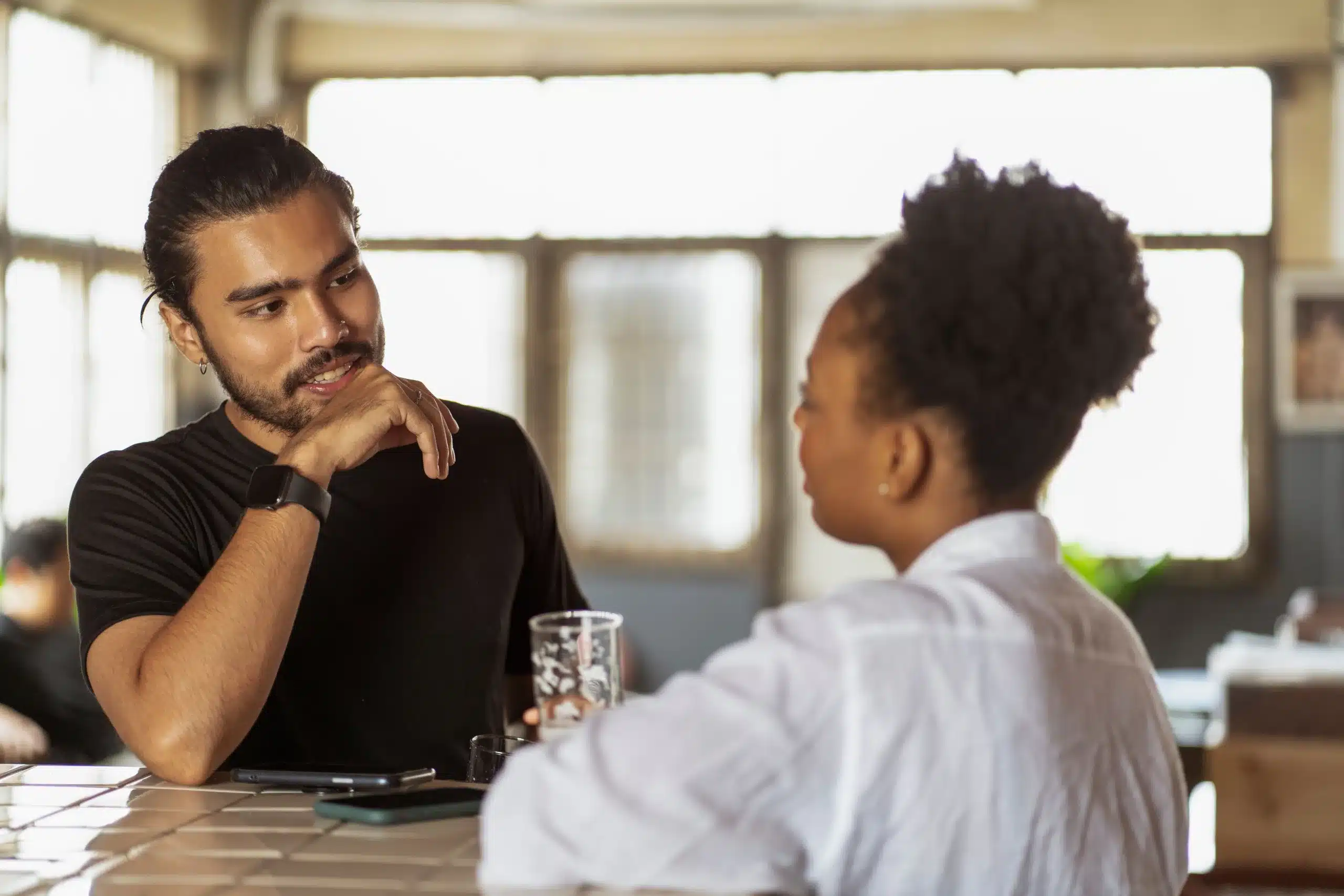Two people talking across a table in a bright room, one listening attentively.