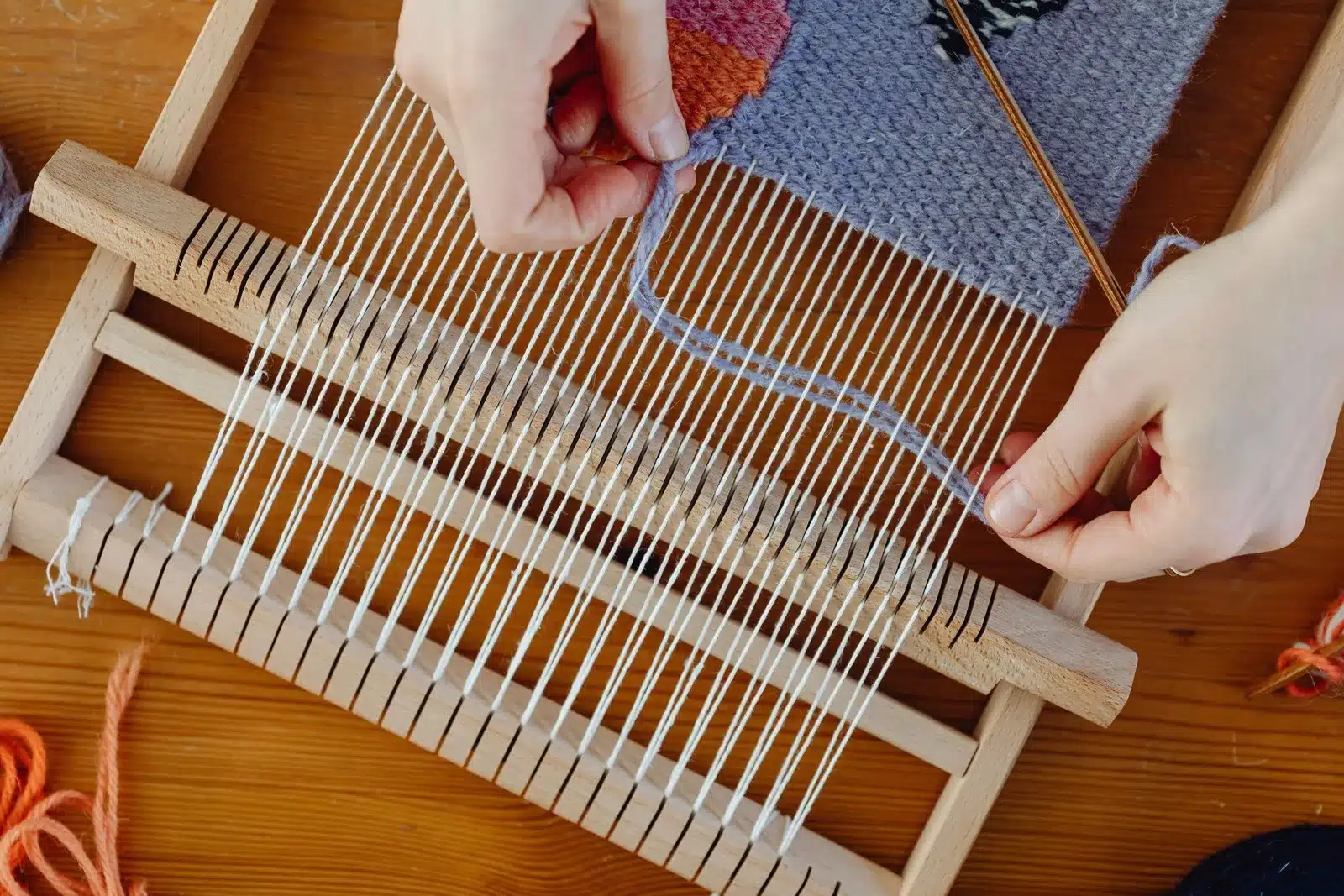Hands working with white threads and a woven blue, rose and rust pattern on a wooden loom.