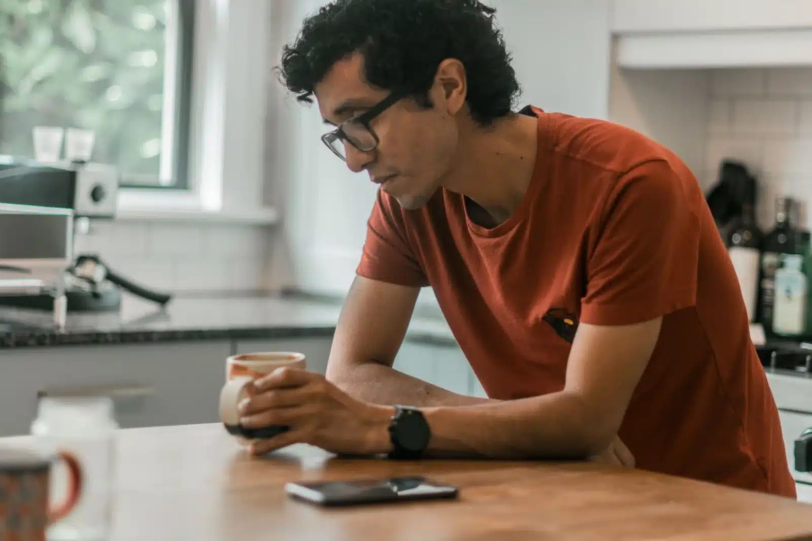 A man leaning on a kitchen counter with a mug of coffee, looking thoughtfully at the phone in front of him.